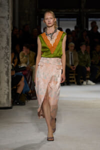 Model walking a runway in a green satin top with orange lace trim and a pale pink floral skirt with a front slit, audience in background.