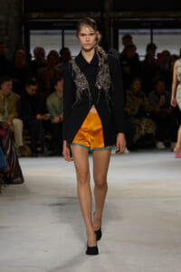 Model walks the runway in a black blazer with silver beaded appliqué draping over bright orange satin shorts with turquoise lace trim.
