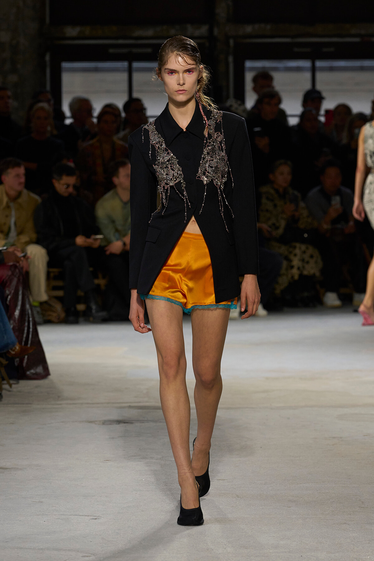 Model walks the runway in a black blazer with silver beaded appliqué draping over bright orange satin shorts with turquoise lace trim.