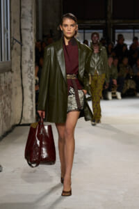 Runway model in an oversized olive leather coat over a burgundy top, patterned shorts, and a large burgundy handbag.
