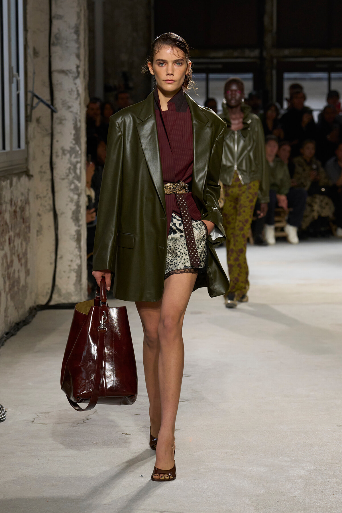 Runway model in an oversized olive leather coat over a burgundy top, patterned shorts, and a large burgundy handbag.