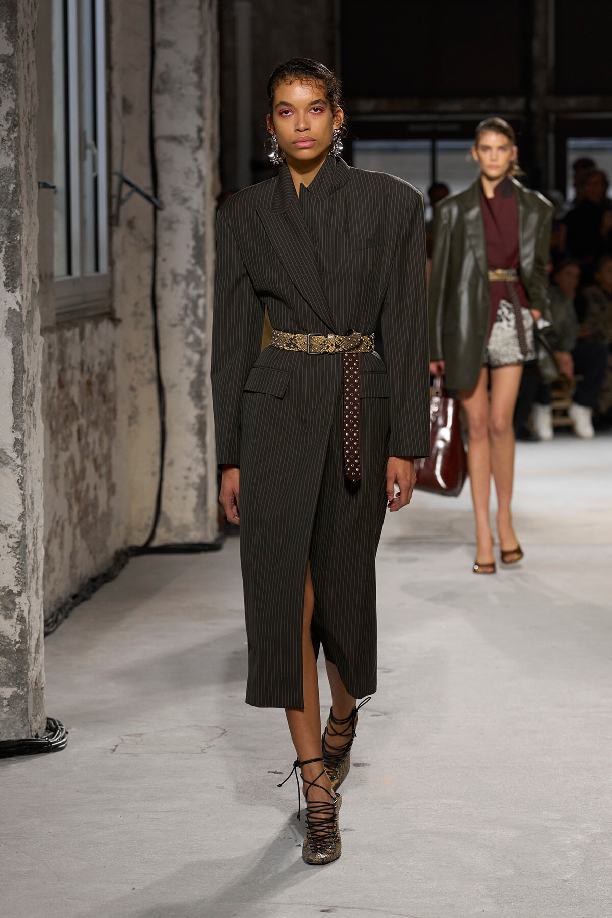 Runway model in a dark pinstripe suit with a wide belt, walking down a gray industrial backstage runway.
