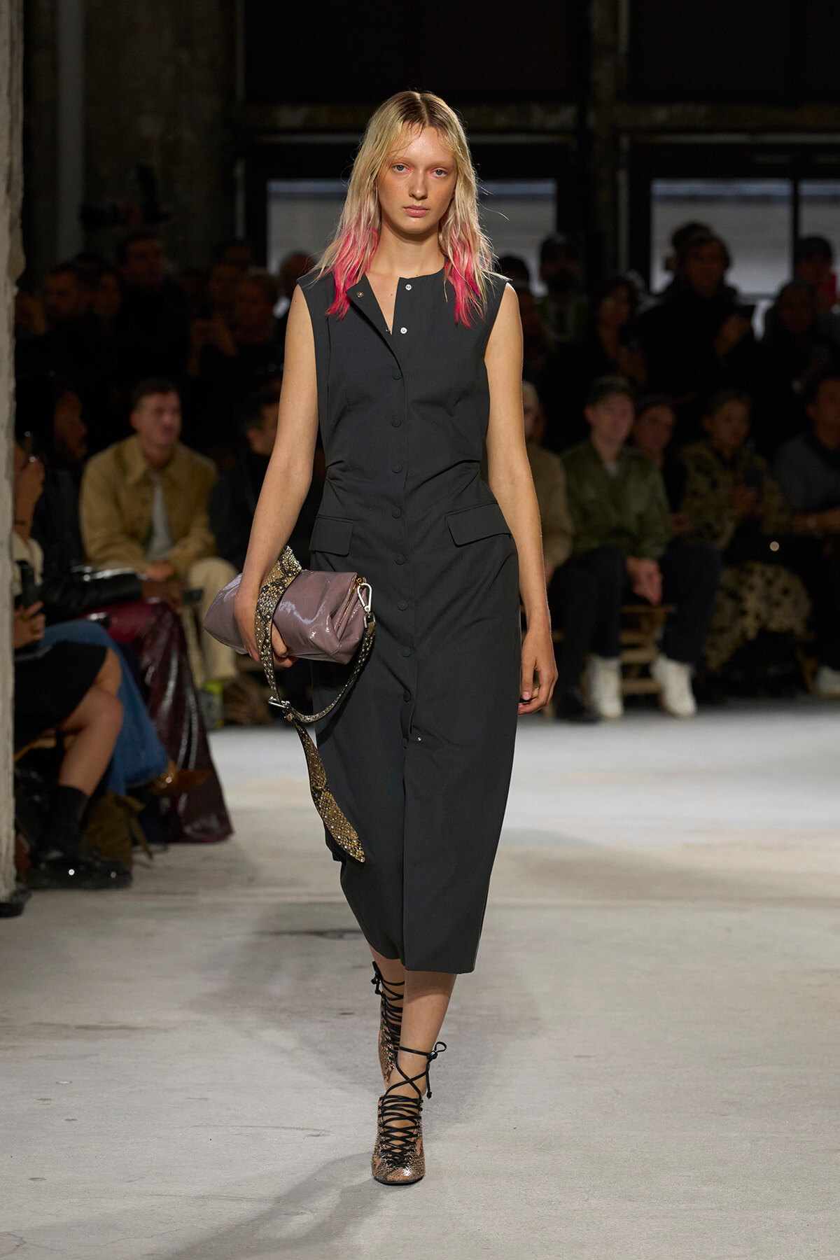 Model on the runway wearing a dark sleeveless button-front jumpsuit with pockets, carrying a mauve clutch and glittery lace-up heels.