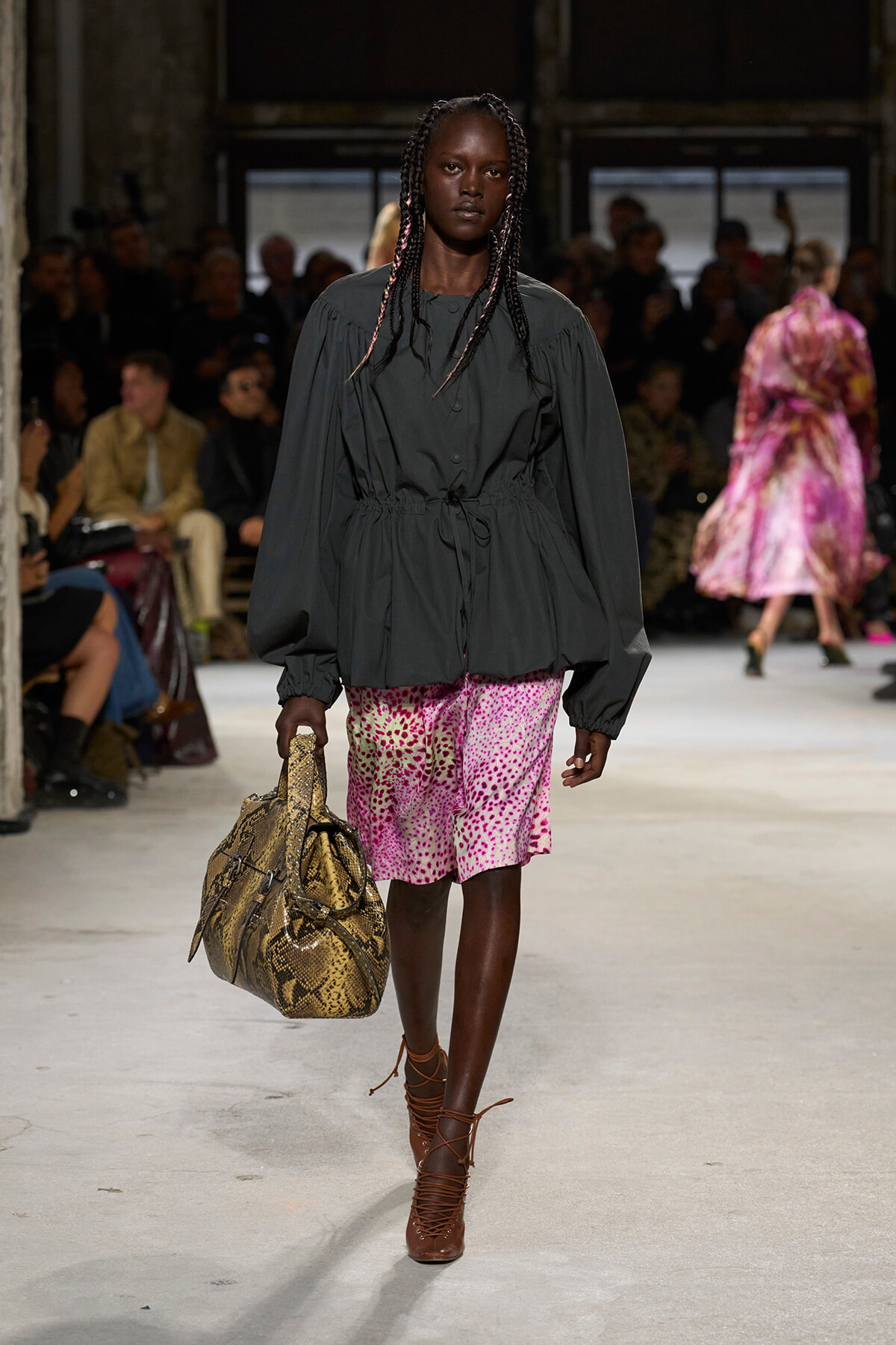 Model on a runway wearing a dark cinched-waist jacket with a pink spotted skirt, brown lace-up heels, and a snakeskin handbag.