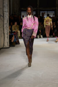 Model walks the runway in a pink spotted blouse tucked into a dark, speckled wrap skirt; industrial venue with seated audience in the background.