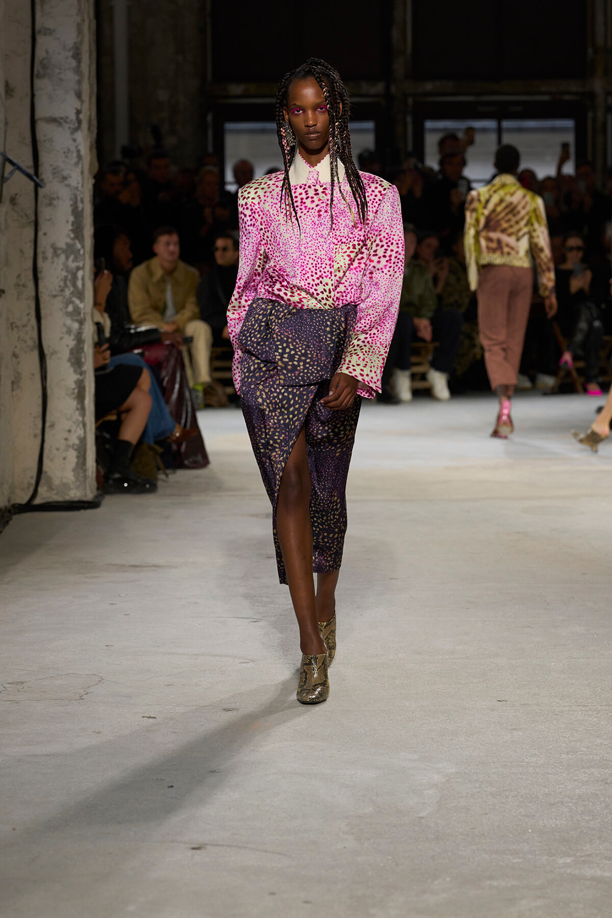 Model walks the runway in a pink spotted blouse tucked into a dark, speckled wrap skirt; industrial venue with seated audience in the background.