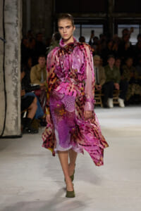 Model walking the runway in a glossy pink and purple abstract-patterned trench coat with a belt, green heels, audience in the background.