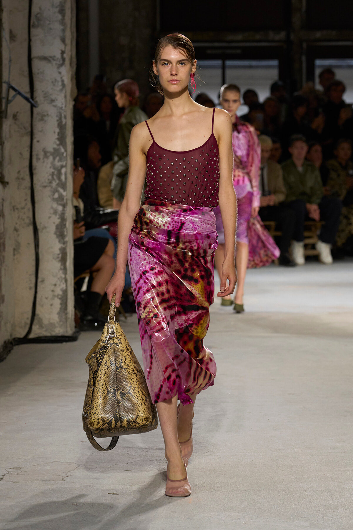 Model on a runway wearing a burgundy studded camisole and a pink-purple tie-dye skirt, carrying a snakeskin bag.