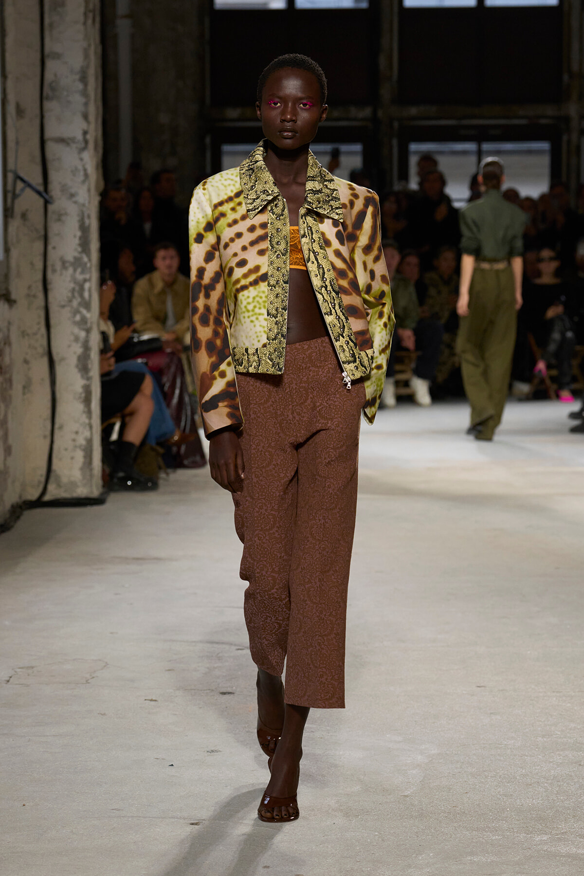 Model walks a fashion runway in a bold animal-print jacket over brown patterned trousers, orange top visible, audience in background.
