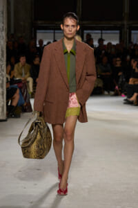 Model on runway wearing a brown patterned oversized blazer over a green top, pink satin shorts with yellow lace, red embellished heels, and a snakeskin bag.