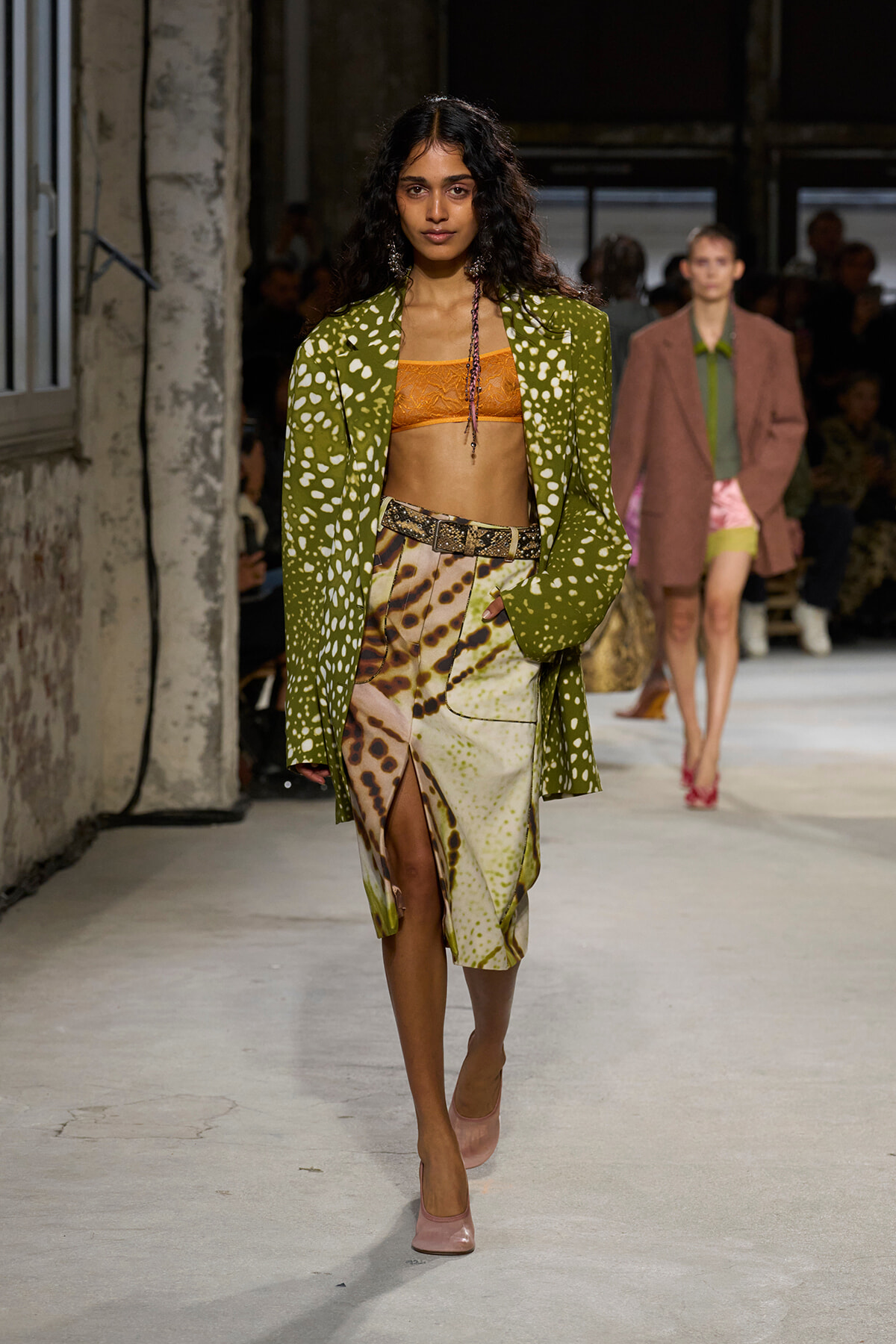 Model walking a runway in a lime green polka-dotted blazer, orange bandeau, animal-print skirt, belt, and pink heels.