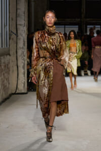 Model walking runway in metallic bronze-brown dress with sheer overlay, high neck, and belt, paired with lace-up metallic heels.