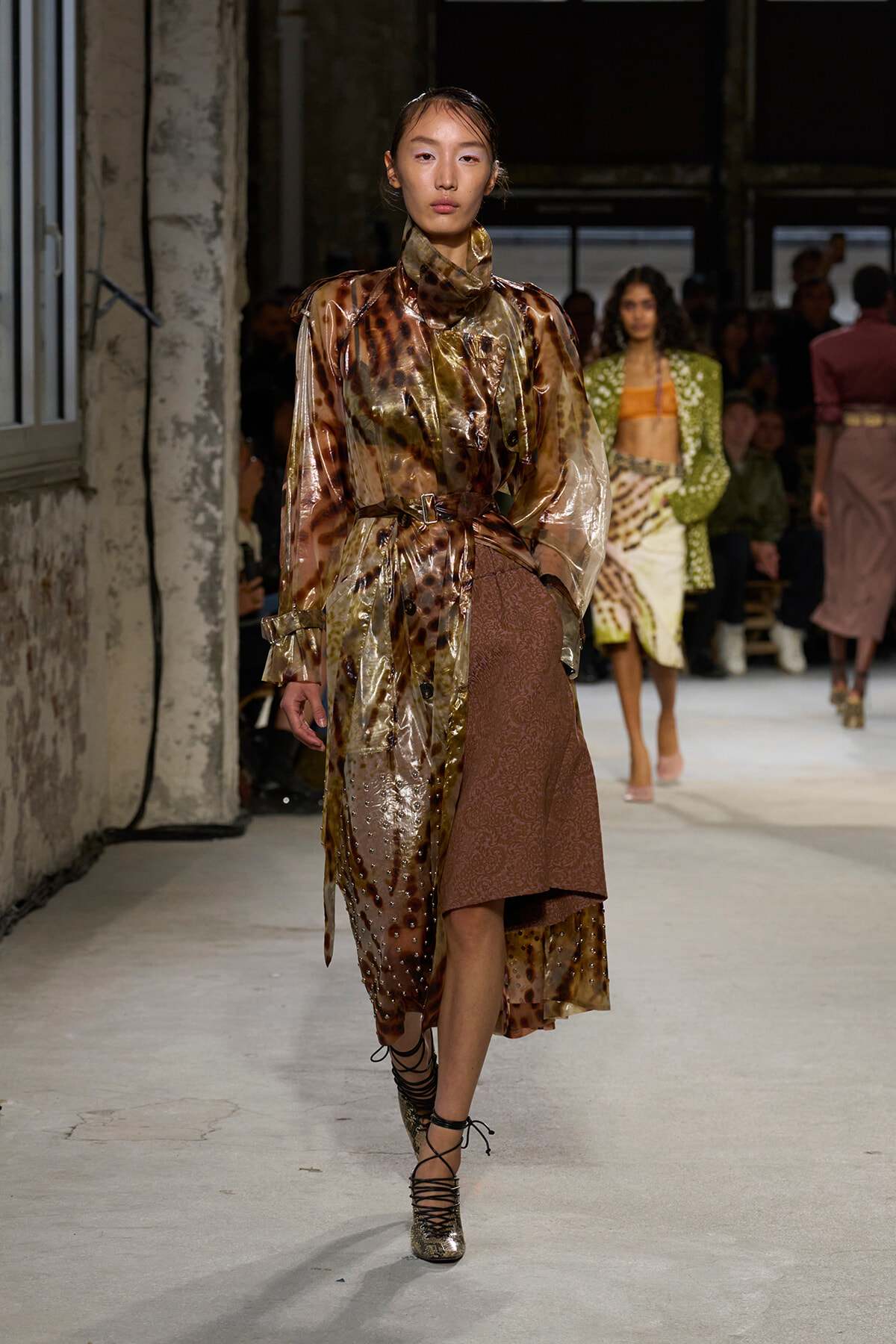 Model walking runway in metallic bronze-brown dress with sheer overlay, high neck, and belt, paired with lace-up metallic heels.