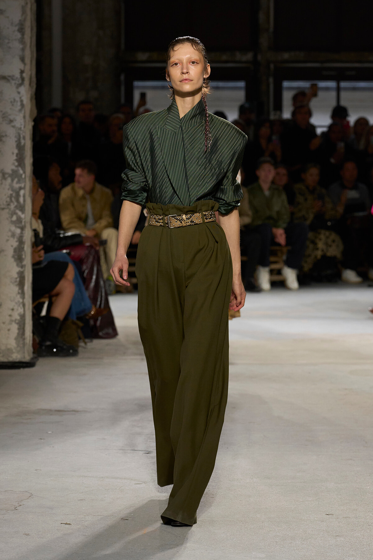 Model on a runway wearing a dark green pinstriped blazer tucked into olive wide-leg pants with a studded belt.