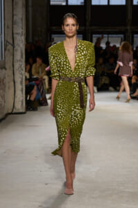 Model walks the runway in a green polka‑dot midi dress with a deep V neckline and a brown belt.