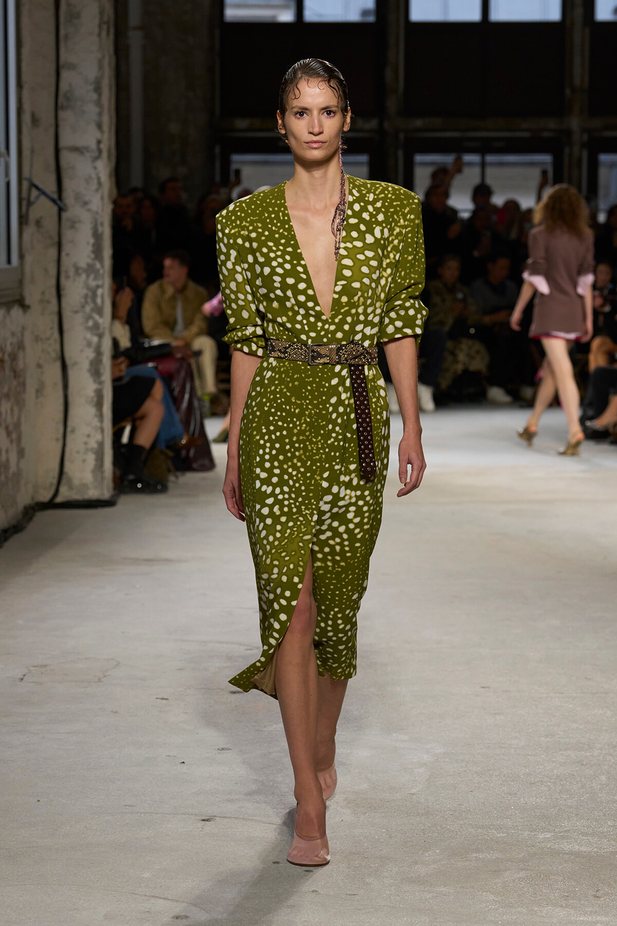 Model walks the runway in a green polka‑dot midi dress with a deep V neckline and a brown belt.