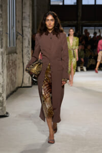 Model on a runway wearing a brown tailored coat over a glossy animal-print dress, carrying a snakeskin clutch.