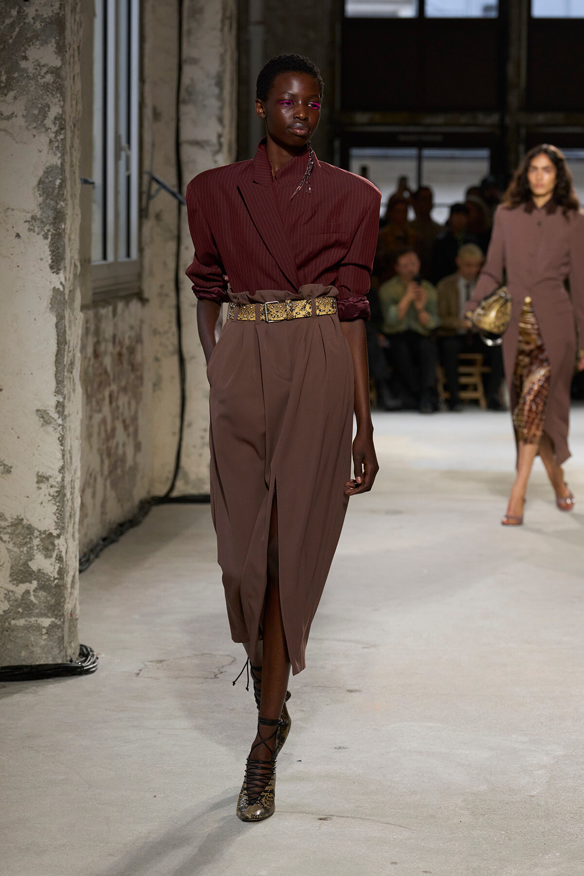 Runway model in a burgundy pinstripe blouse tucked into a brown draped skirt with a gold belt walking the catwalk.