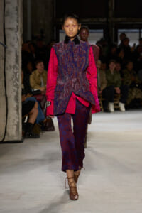 Model walks the runway in a pink and dark blue patterned suit with bright pink sleeves, brown lace-up heels, and an attentive audience in the background.