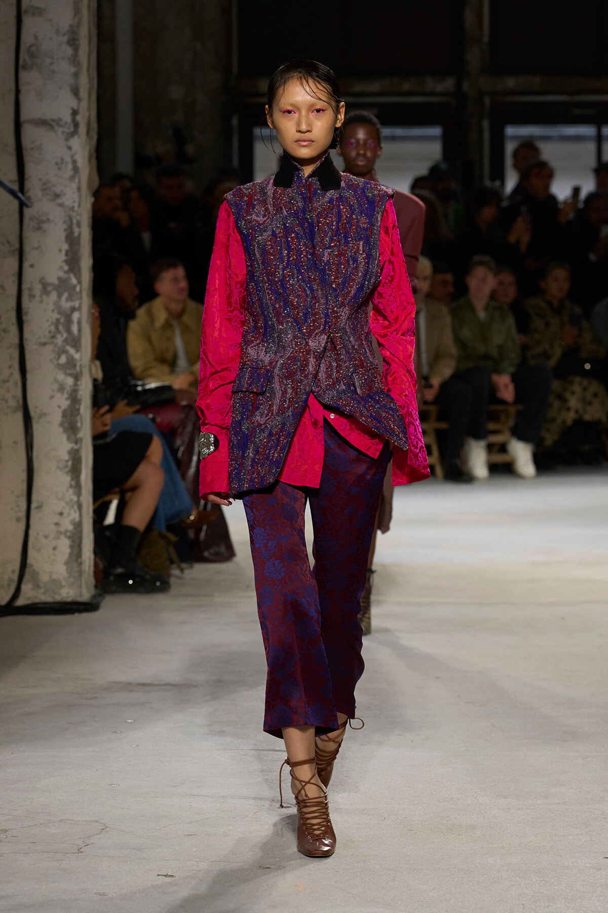 Model walks the runway in a pink and dark blue patterned suit with bright pink sleeves, brown lace-up heels, and an attentive audience in the background.
