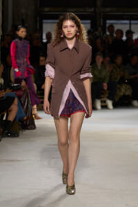 Model walks the runway in a taupe oversized shirt layered over a pink blouse, paired with patterned shorts and snakeskin heels, audience in background.