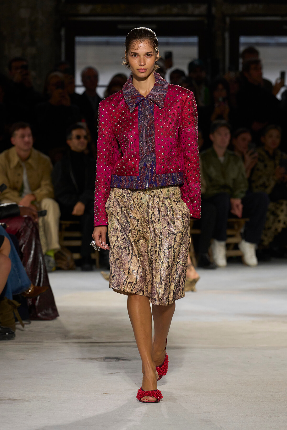 Model walking a runway in a bright pink studded jacket over a patterned blouse and a snakeskin midi skirt, red heeled sandals.