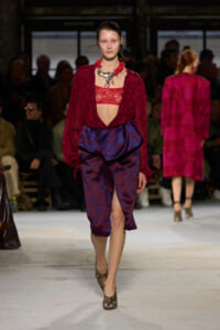 Model walking the runway in a burgundy lace blouse and voluminous blue patterned skirt with a deep V at the midriff, statement necklace, and snakeskin heels.