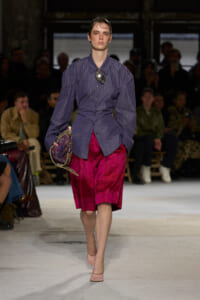 Model walks a runway in a striped purple button-down shirt with a large statement necklace and a magenta velvet knee-length skirt, carrying a patterned bag.