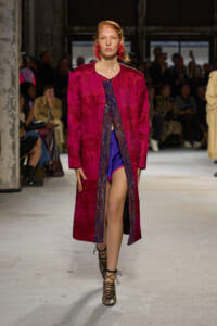 Model walks the runway in a bright magenta textured coat over a blue outfit, paired with bold red earrings.
