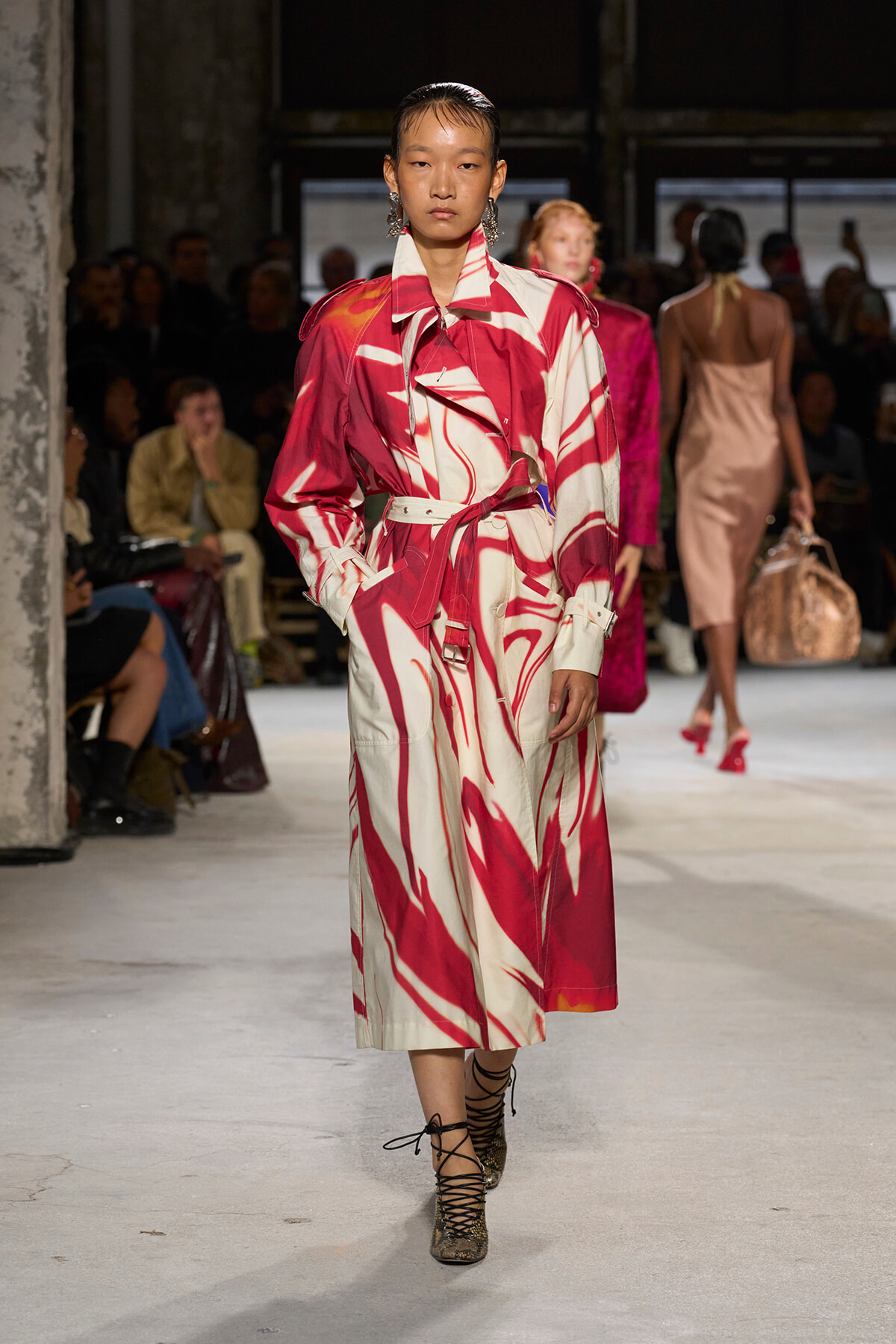 Model walking a runway in a red-and-cream abstract print trench dress with belt, statement earrings, and lace-up sandals.