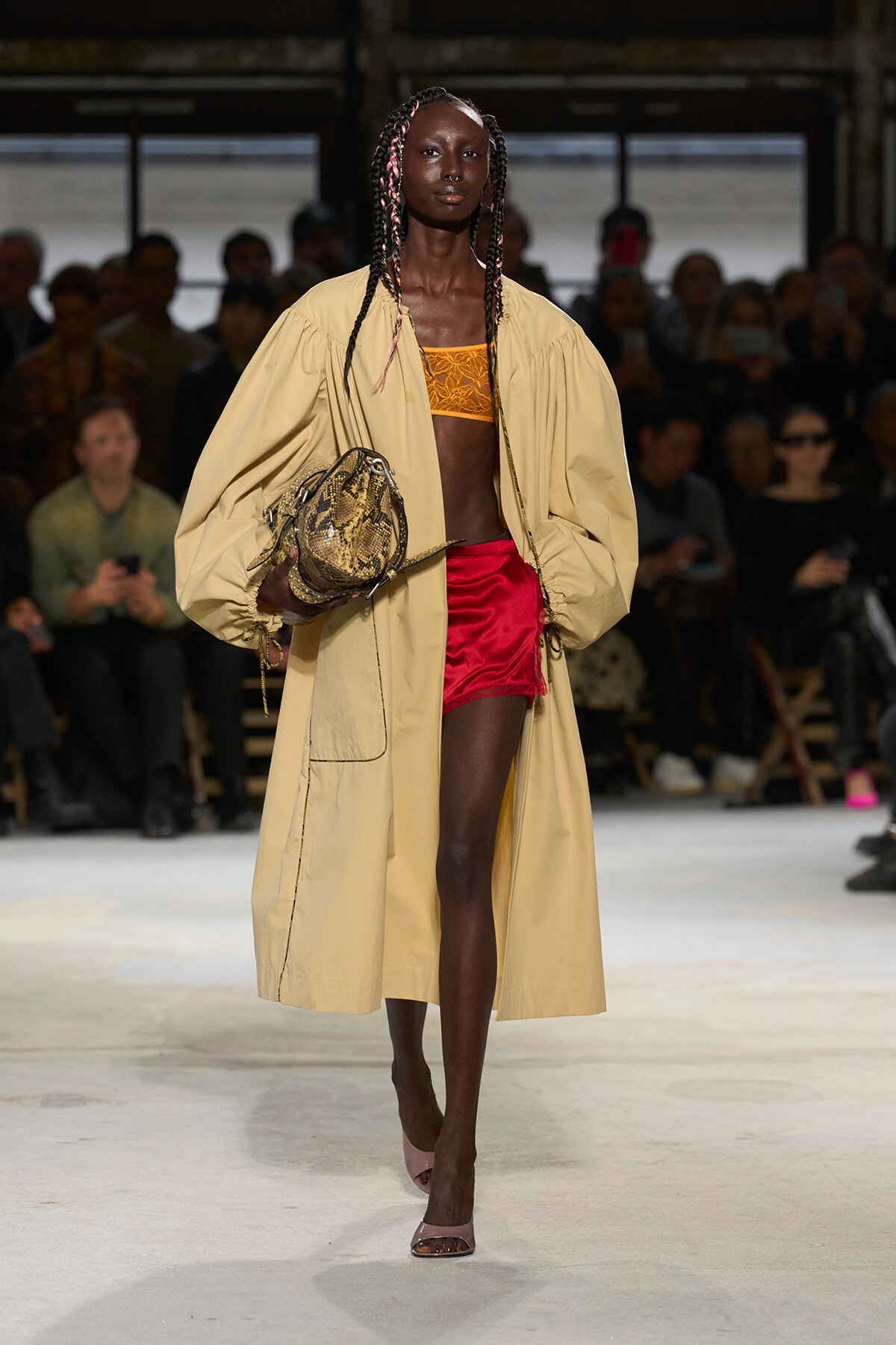 Model on a runway wearing a beige oversized trench, orange bralette, and red satin shorts, carrying a snakeskin handbag.