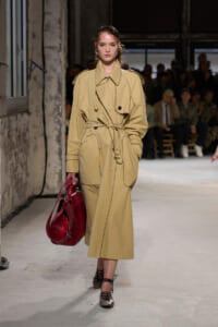 Model on a runway wearing a belted beige trench coat, carrying a red textured handbag.