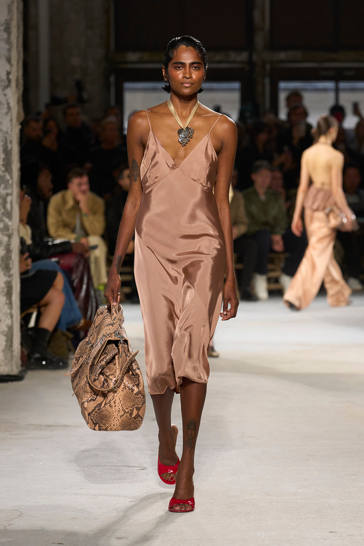 Model walks a runway in a blush satin slip dress, red open-toe heels, and a snakeskin bag during a fashion show