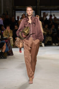 Model walks a fashion runway in a dusty-rose velvet jacket with a ruffled front and matching satin pants, carrying a metallic gold snakeskin handbag.