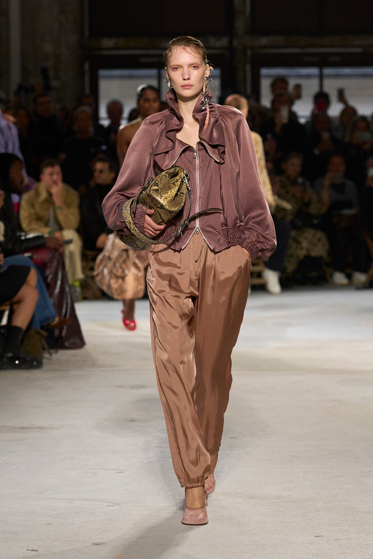 Model walks a fashion runway in a dusty-rose velvet jacket with a ruffled front and matching satin pants, carrying a metallic gold snakeskin handbag.
