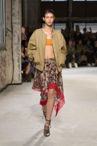 Model walking a runway in a beige bomber jacket over an orange bralette and a patterned, red-trimmed skirt with lace hem.