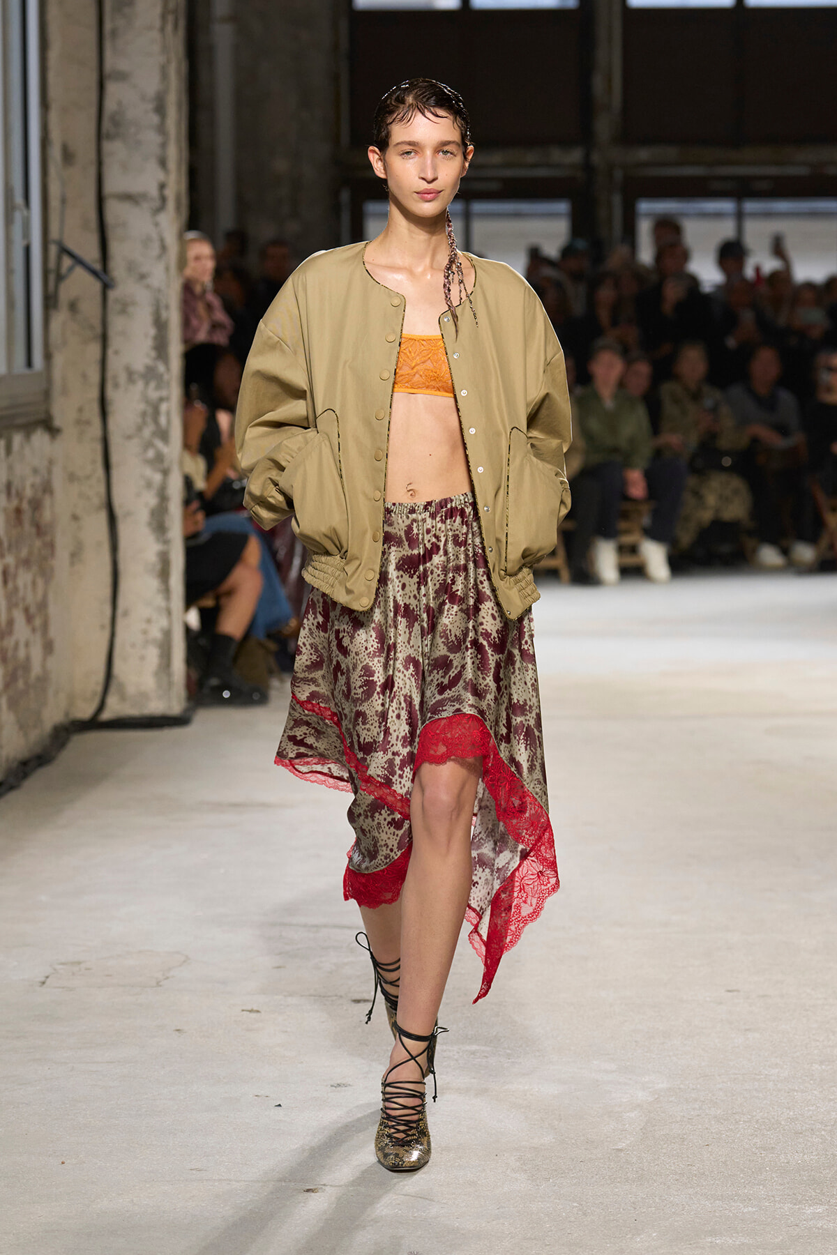 Model walking a runway in a beige bomber jacket over an orange bralette and a patterned, red-trimmed skirt with lace hem.