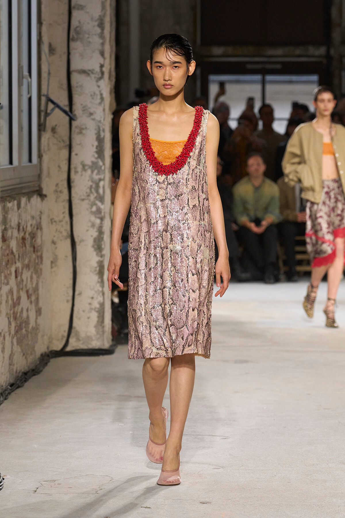 Model walking the runway in a pink snakeskin slip dress with orange underlayer and a red beaded neckline; audience in background.
