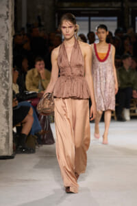 Model walking a fashion runway in a pink satin halter peplum top and wide-leg pants, holding a small metallic bag; braided hair, focused expression.