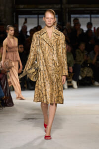 Model walks the runway wearing a gold snake-print coat and carrying a metallic bag, paired with red sandals.