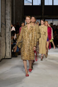 Models walk a runway at a fashion show, lead model wearing a gold snakeskin coat and red sandals.