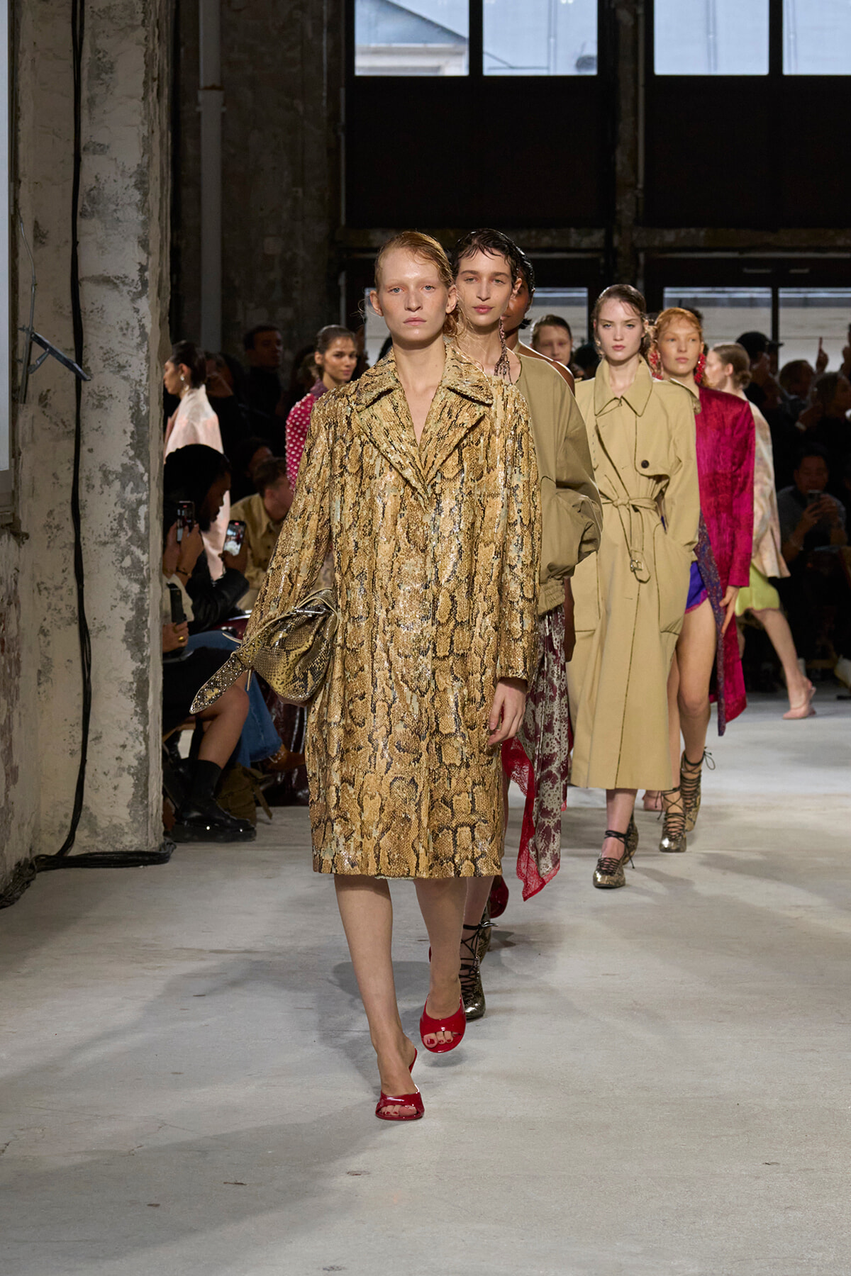 Models walk a runway at a fashion show, lead model wearing a gold snakeskin coat and red sandals.