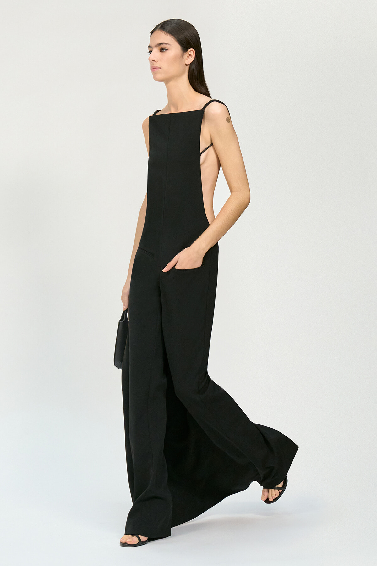 Model wearing a black sleeveless jumpsuit with side cutouts and wide legs, walking forward in sandals.