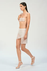 Model walking in a beige two-piece outfit: bandeau top and asymmetrical wrap skirt, white heeled sandals.