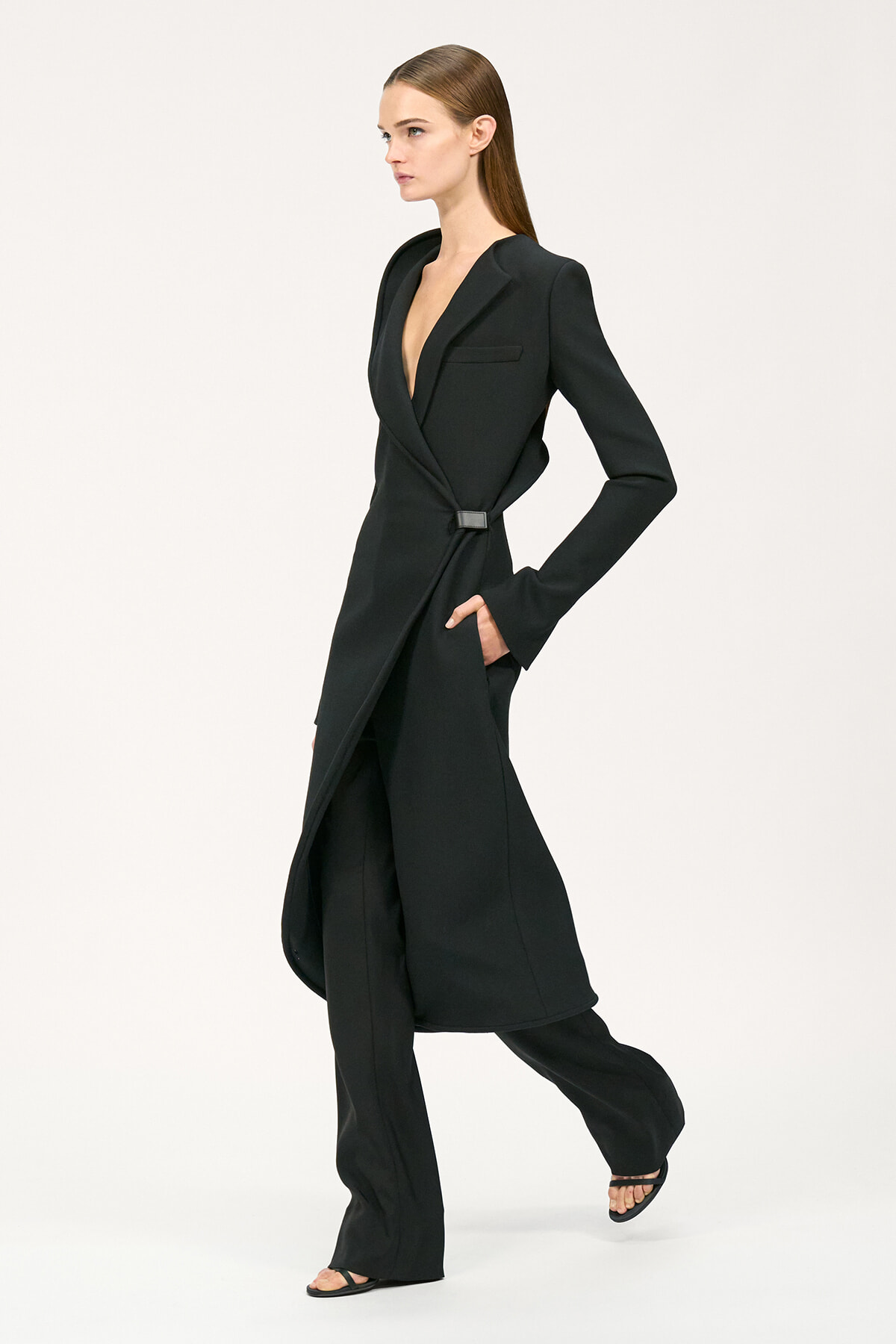 Model wearing a black long belted blazer over matching trousers, shown in profile in a studio setting.