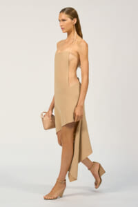 Model wearing a nude strapless beige dress with a high-low hem, carrying a small beige handbag and wearing nude sandals.