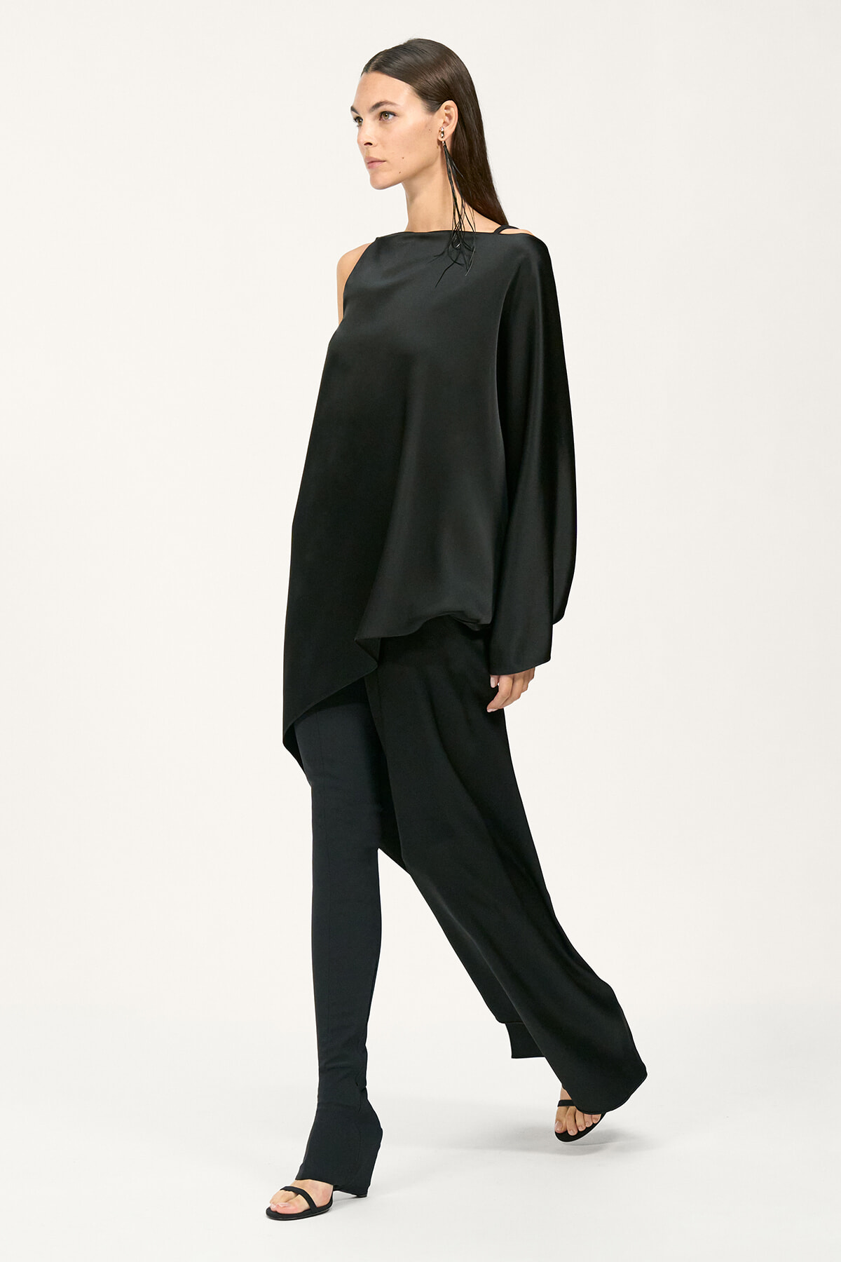 Woman in an all-black, draped one-shoulder top with wide-leg pants and open-toe sandals, posing against a light background.