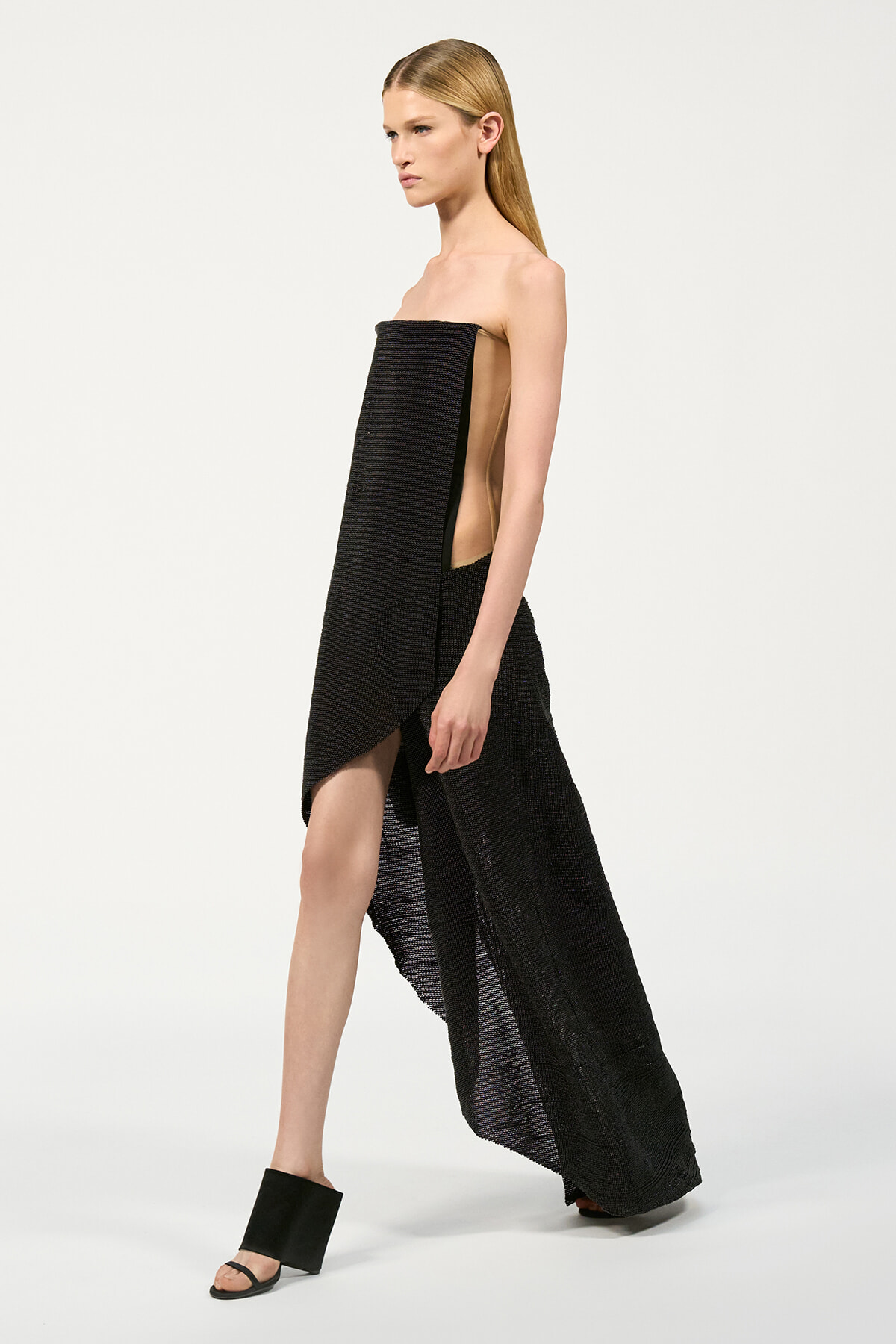 Model in a strapless black textured gown with a long draped overlay and high side slit, standing in profile.