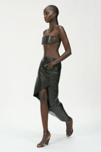 Side-profile of a model wearing a black leather bandeau top and an asymmetrical wrap skirt with heeled sandals