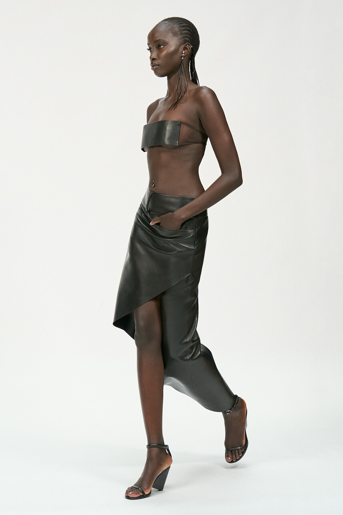 Side-profile of a model wearing a black leather bandeau top and an asymmetrical wrap skirt with heeled sandals